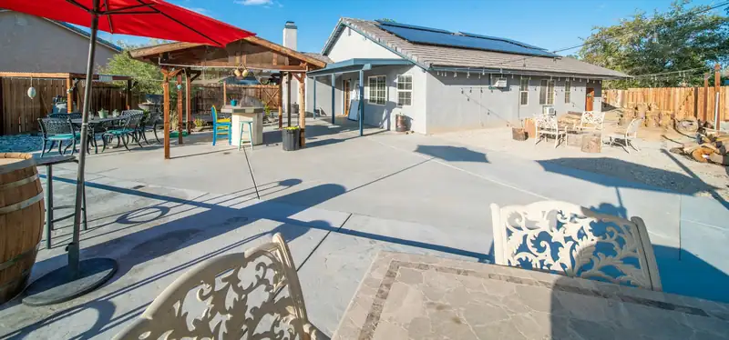 Large backyard concrete patio with umbrellas and a pergola behind a single-story home