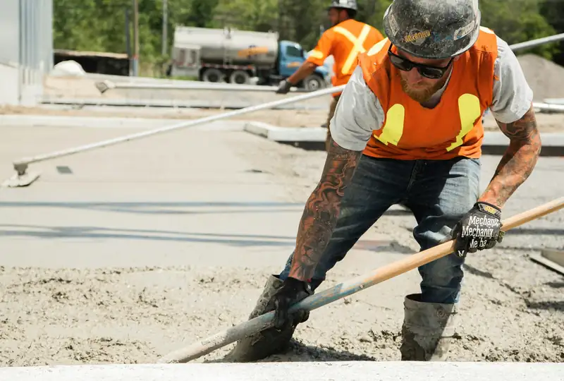 Worker in a hard hat and orange vest spreading fresh concrete on a sunny site