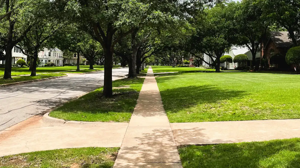 Straight residential concrete sidewalk under shade trees crossing a curb approach