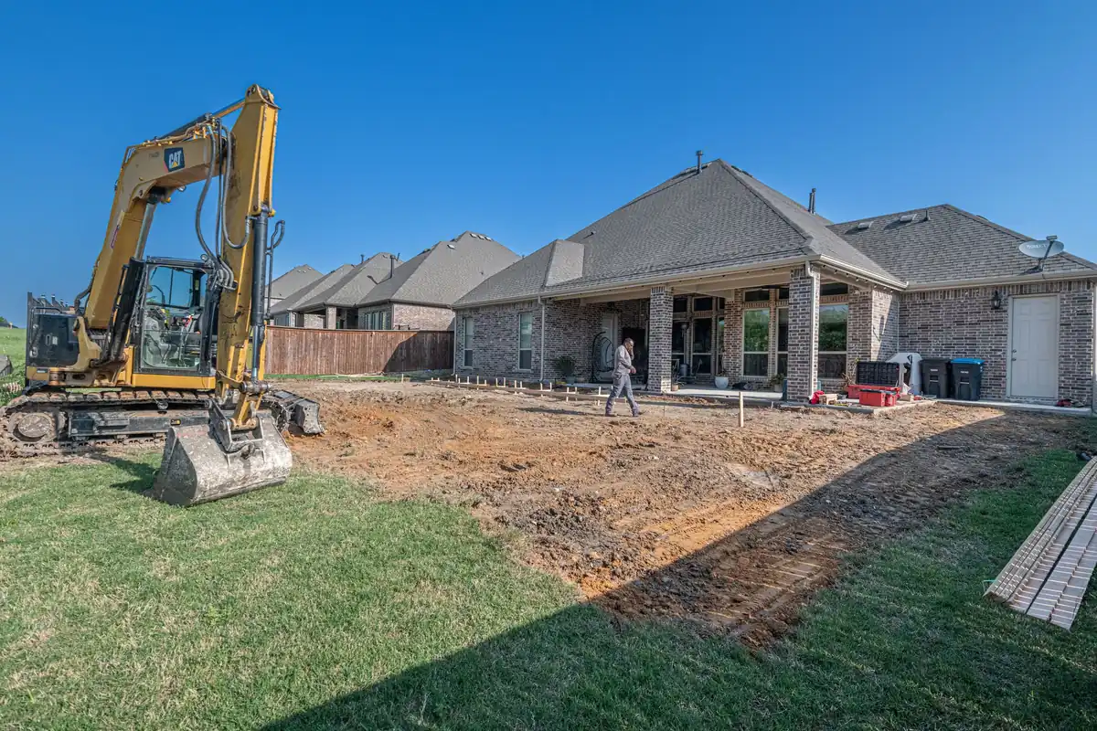 Excavator grading a backyard behind a brick home before a new concrete patio